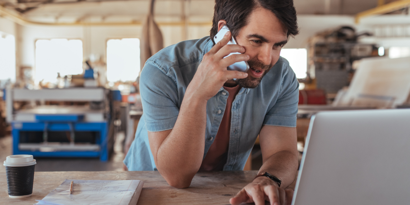 Mitarbeiter eines Handwerksbetriebs telefoniert und arbeitet gleichzeitig am Laptop – Symbol für zuverlässige Business-Telefonie und moderne Kommunikation über Glasfaser.