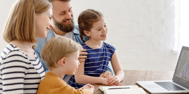 Eine lächelnde Familie sitzt gemeinsam am Tisch und schaut auf einen Laptop.